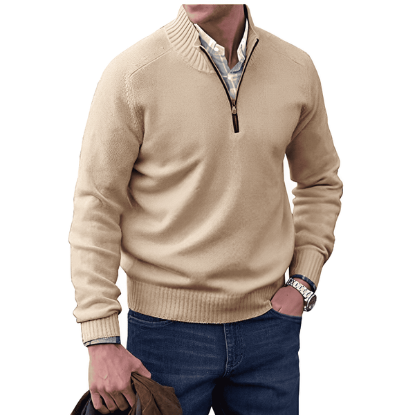 Homme portant un pull beige à col zippé, chemise blanche, jeans bleu, montre argentée, tenant une veste marron. Mode masculine élégante et décontractée.