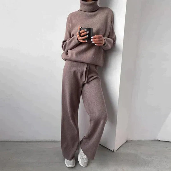 Femme en ensemble pull et pantalon en laine beige, tenant une tasse, appuyée contre un mur blanc. Mode décontractée, tenue confortable automne-hiver.