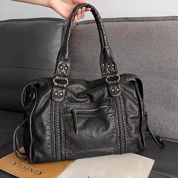 Sac à main en cuir noir texturé avec poignées robustes, poche zippée avant, posé sur un canapé gris. Accessoire mode élégant et pratique.