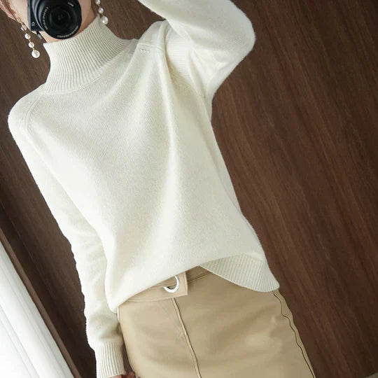 Femme portant un pull blanc à col roulé et une jupe beige, posant devant un mur en bois. Mode automne-hiver, tenue élégante et chic.