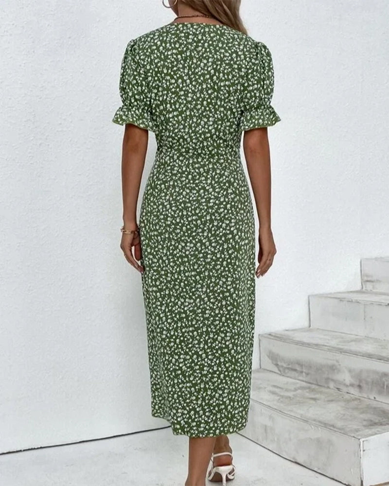 Robe longue verte à motifs floraux blancs, manches courtes bouffantes, portée par une femme sur fond de mur blanc et escaliers. Mode féminine élégante.