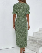 Robe longue verte à motifs floraux blancs, manches courtes bouffantes, portée par une femme sur fond de mur blanc et escaliers. Mode féminine élégante.