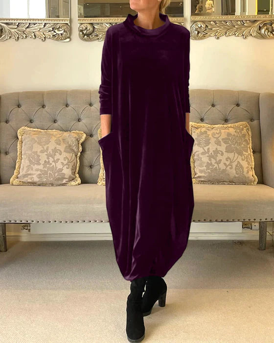 Robe longue en velours violet avec poches, portée par une femme debout devant un canapé élégant, décoré de coussins à motifs floraux.