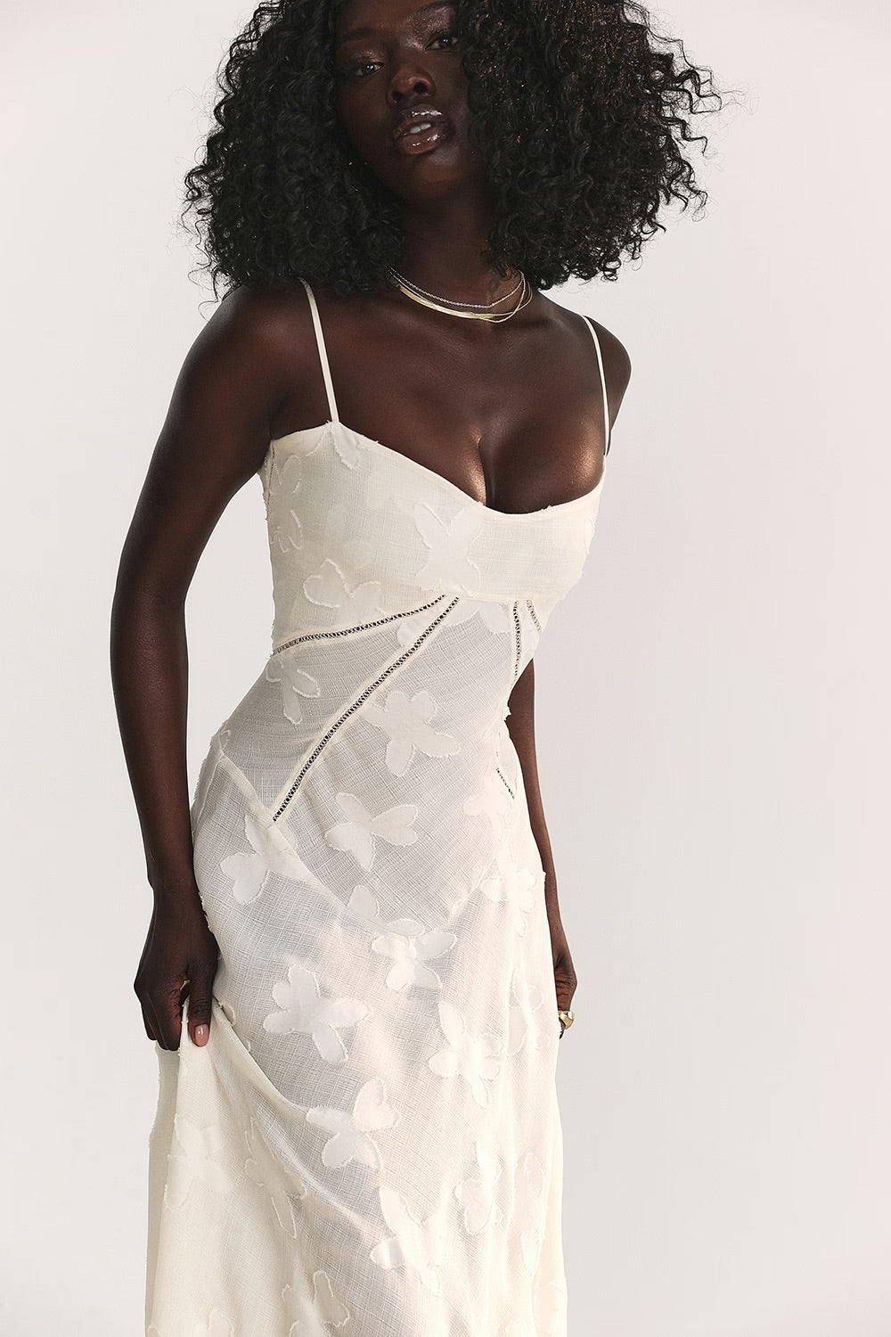 Femme portant une robe blanche élégante avec motifs floraux, bretelles fines et col en cœur, posant sur fond neutre. Mode féminine chic et tendance.