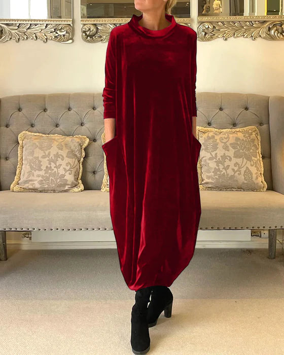 Robe longue en velours rouge avec poches, portée par une personne debout devant un canapé élégant. Mode automne-hiver, style chic et confortable.