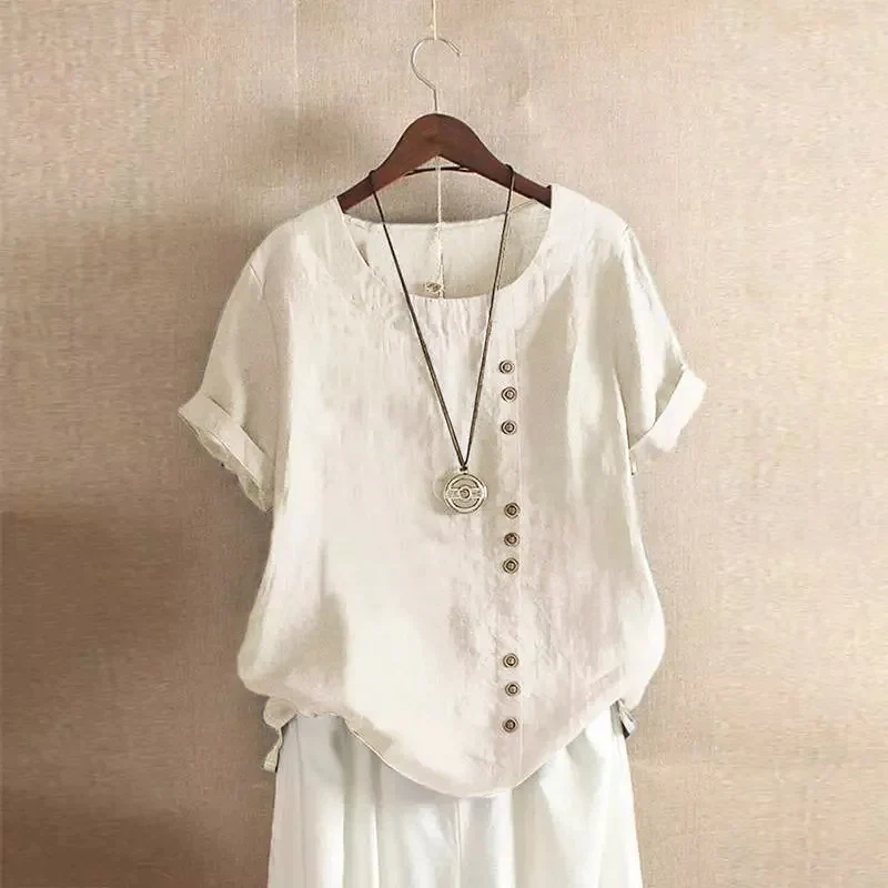 Blouse en lin blanc avec boutons décoratifs, manches courtes retroussées, col rond, suspendue sur un cintre en bois. Mode féminine élégante.