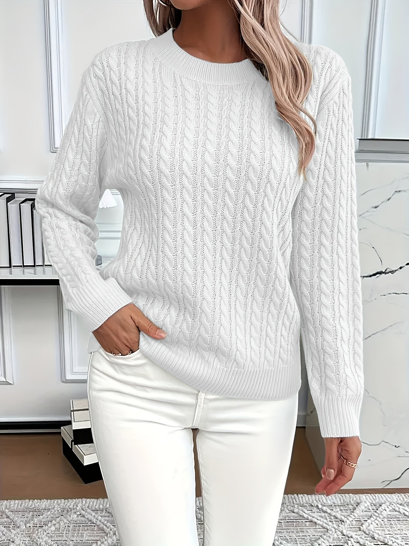 Femme portant un pull torsadé blanc élégant, manches longues, assorti à un pantalon blanc, posant dans un intérieur moderne et lumineux.