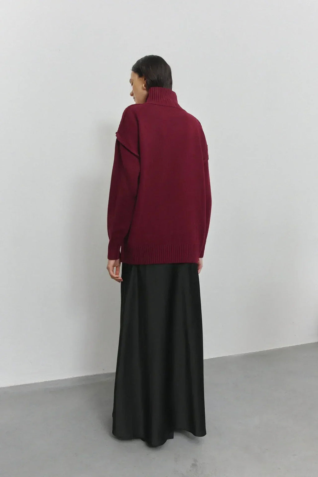 Femme portant un pull bordeaux à col roulé et une jupe longue noire, debout contre un mur blanc. Mode automne-hiver, style décontracté élégant.