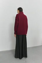 Femme portant un pull bordeaux à col roulé et une jupe longue noire, debout contre un mur blanc. Mode automne-hiver, style décontracté élégant.