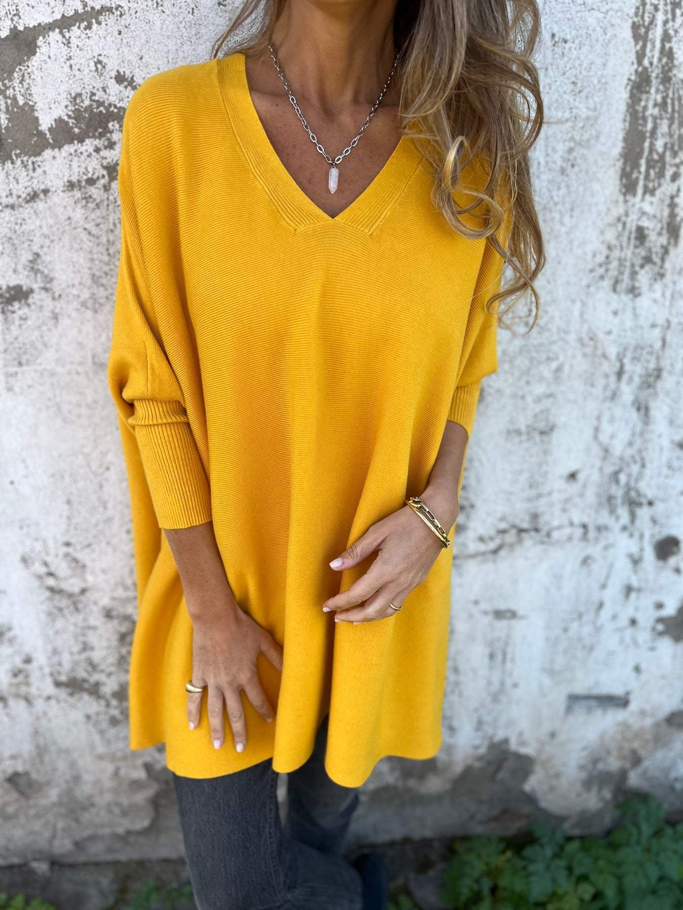 Femme portant un pull oversize jaune vif, col en V, posant devant un mur texturé. Mode automne, style décontracté, tendance féminine.