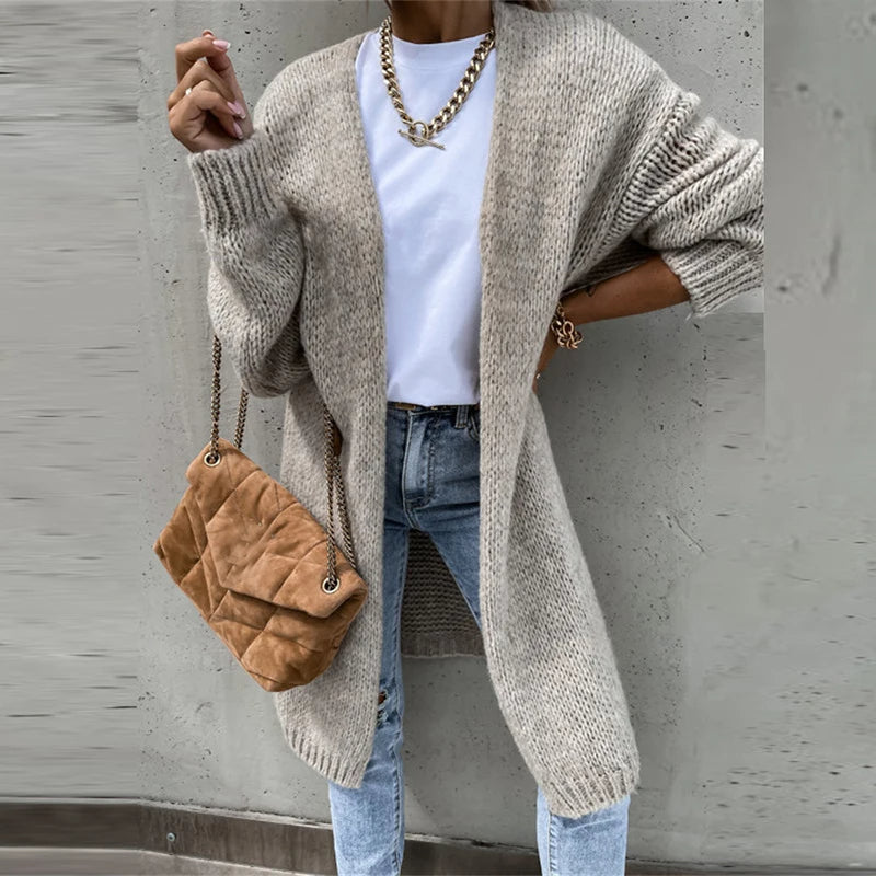 Femme portant un long cardigan gris, t-shirt blanc, jean bleu, sac à main beige matelassé, chaîne dorée, style décontracté chic.