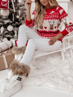 Femme en pull de Noël rouge avec motifs de rennes, assise près d'un sapin décoré, portant des bottes blanches à fourrure, ambiance festive.