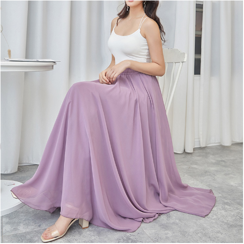 Femme en débardeur blanc et jupe longue violette assise sur une chaise, intérieur moderne, rideaux blancs, sol gris. Mode élégante et décontractée.