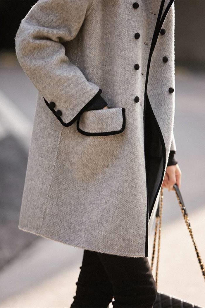 Manteau gris élégant avec boutons noirs, poches à rabat, porté par une personne tenant un sac à main chaîne dorée. Mode automne-hiver chic.