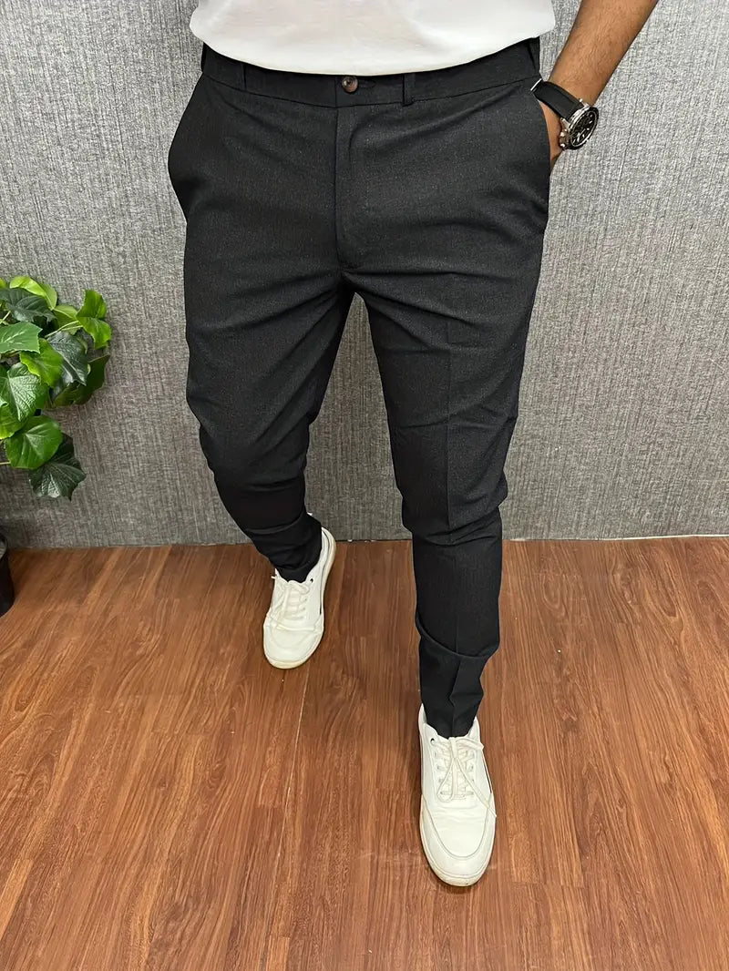 Homme portant un pantalon noir élégant, chaussures blanches, montre noire, sur sol en bois avec plante verte en arrière-plan. Mode masculine chic.