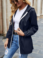 Femme portant un imperméable bleu marine, t-shirt blanc et jeans déchirés, souriante, lunettes de soleil, chaîne dorée, rue pavée en arrière-plan.