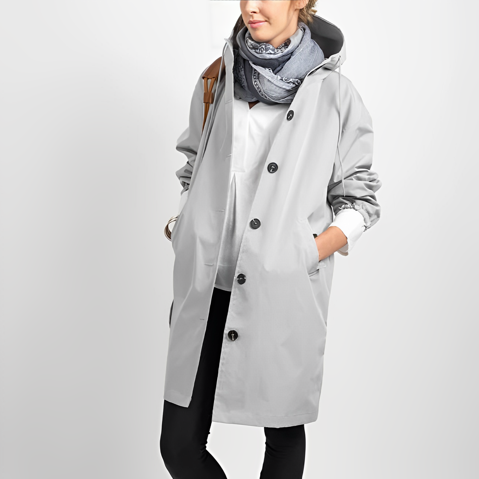 Femme portant un manteau imperméable gris avec capuche, écharpe bleue, pantalon noir, posant contre un fond blanc. Mode automne, style décontracté.