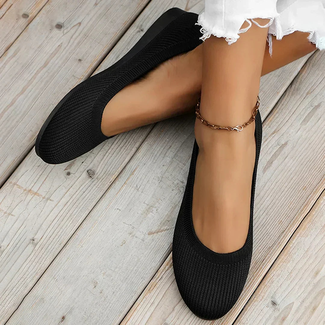Ballerines noires élégantes sur pieds féminins, portées avec un jean blanc effiloché, posées sur un sol en bois. Mode féminine, chaussures confortables.