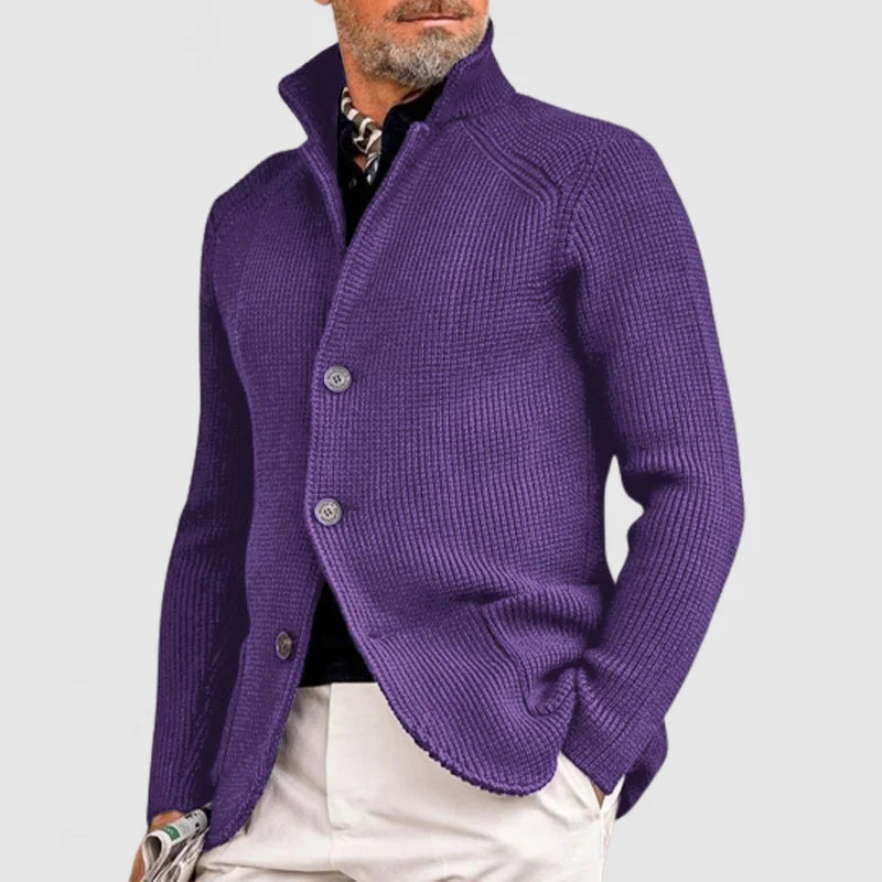 Homme portant un cardigan violet élégant, col relevé, boutons visibles, sur chemise noire et pantalon beige. Mode masculine chic et moderne.