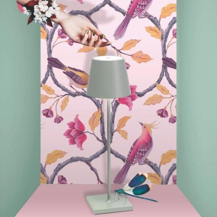 Lampe moderne grise sur fond de papier peint rose avec motifs d'oiseaux et fleurs colorés, décorée d'une libellule bleue et d'une main florale.