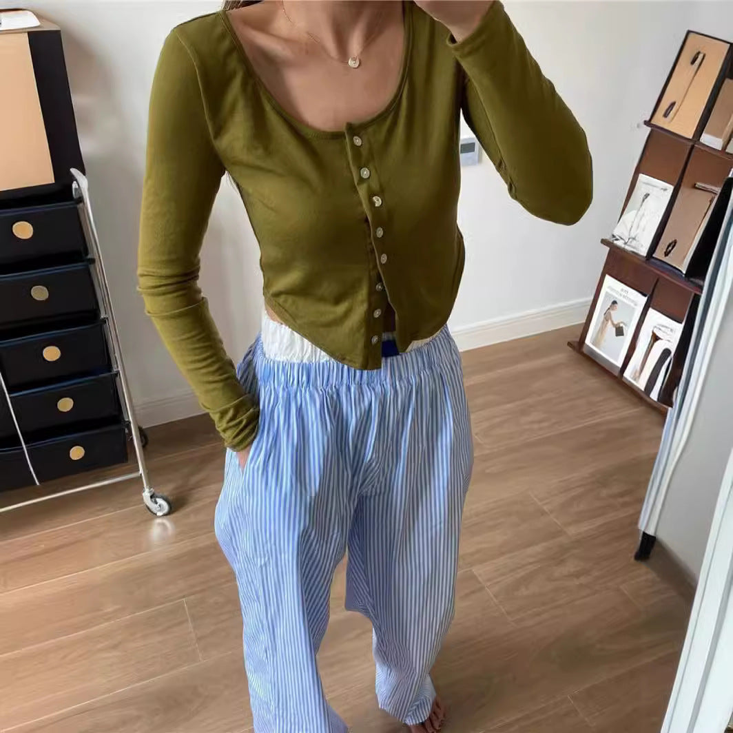 Femme portant un cardigan vert olive et un pantalon rayé bleu et blanc, debout sur un parquet en bois, avec des meubles et cadres en arrière-plan.