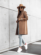 Femme élégante en manteau camel, chapeau assorti, lunettes de soleil, jeans slim et baskets blanches, marchant devant un mur moderne en béton.