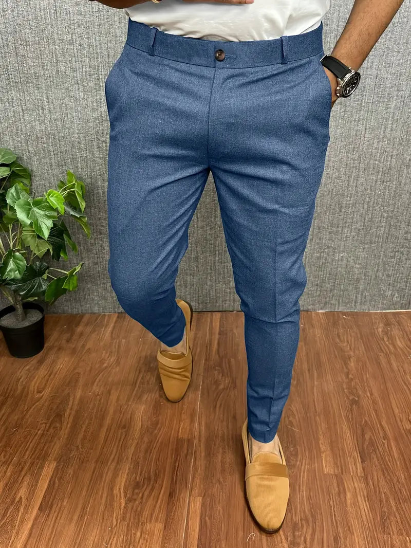 Homme portant un pantalon bleu élégant, chaussures marron clair, montre noire, sur sol en bois avec plante verte en arrière-plan. Mode masculine chic.