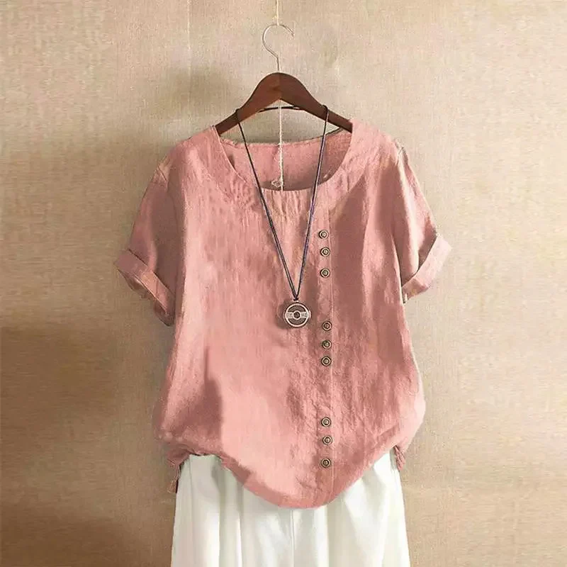 Blouse rose en lin avec boutons asymétriques, manches courtes retroussées, suspendue sur un cintre en bois, fond beige. Mode féminine élégante.