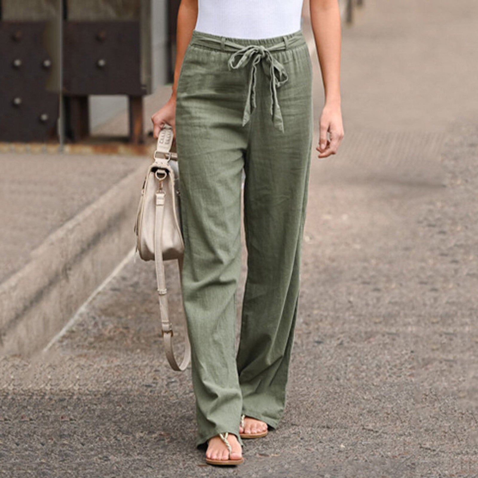 Femme portant un pantalon large vert kaki en lin avec ceinture, sac à main beige, sandales, sur un trottoir urbain. Mode décontractée chic.