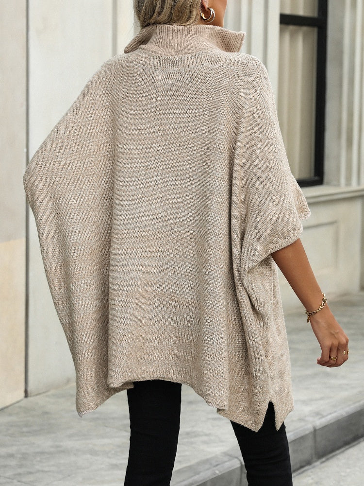 Femme portant un poncho beige en tricot à col roulé, style décontracté, devant un bâtiment moderne. Mode automne-hiver, tendance élégante.
