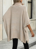 Femme portant un poncho beige en tricot à col roulé, style décontracté, devant un bâtiment moderne. Mode automne-hiver, tendance élégante.