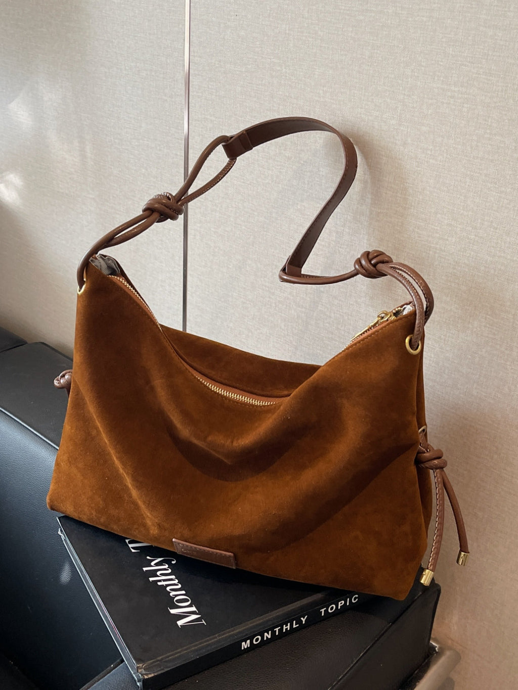 Sac à main en daim marron avec bandoulière en cuir, posé sur un livre noir. Élégant et moderne, idéal pour la mode automne-hiver.