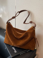 Sac à main en daim marron avec bandoulière en cuir, posé sur un livre noir. Élégant et moderne, idéal pour la mode automne-hiver.