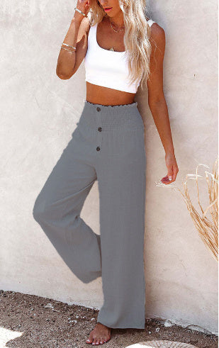 Femme en débardeur blanc et pantalon large gris, posant contre un mur beige. Mode estivale, tenue décontractée, style chic et tendance.