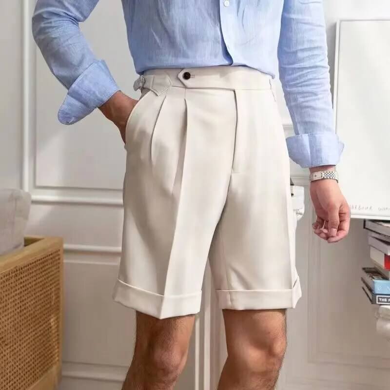 Homme en chemise bleue et short beige élégant, debout dans un intérieur moderne. Mode masculine, tenue estivale chic, style décontracté.