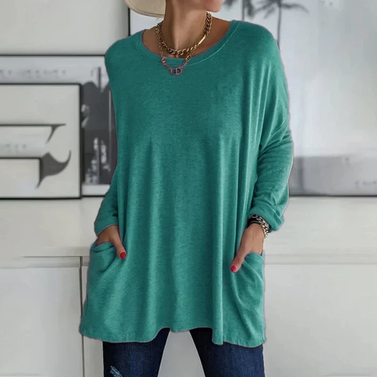 Femme portant un pull vert ample avec poches, collier doré et chapeau beige, posant devant un mur décoré. Mode décontractée, tendance automne.