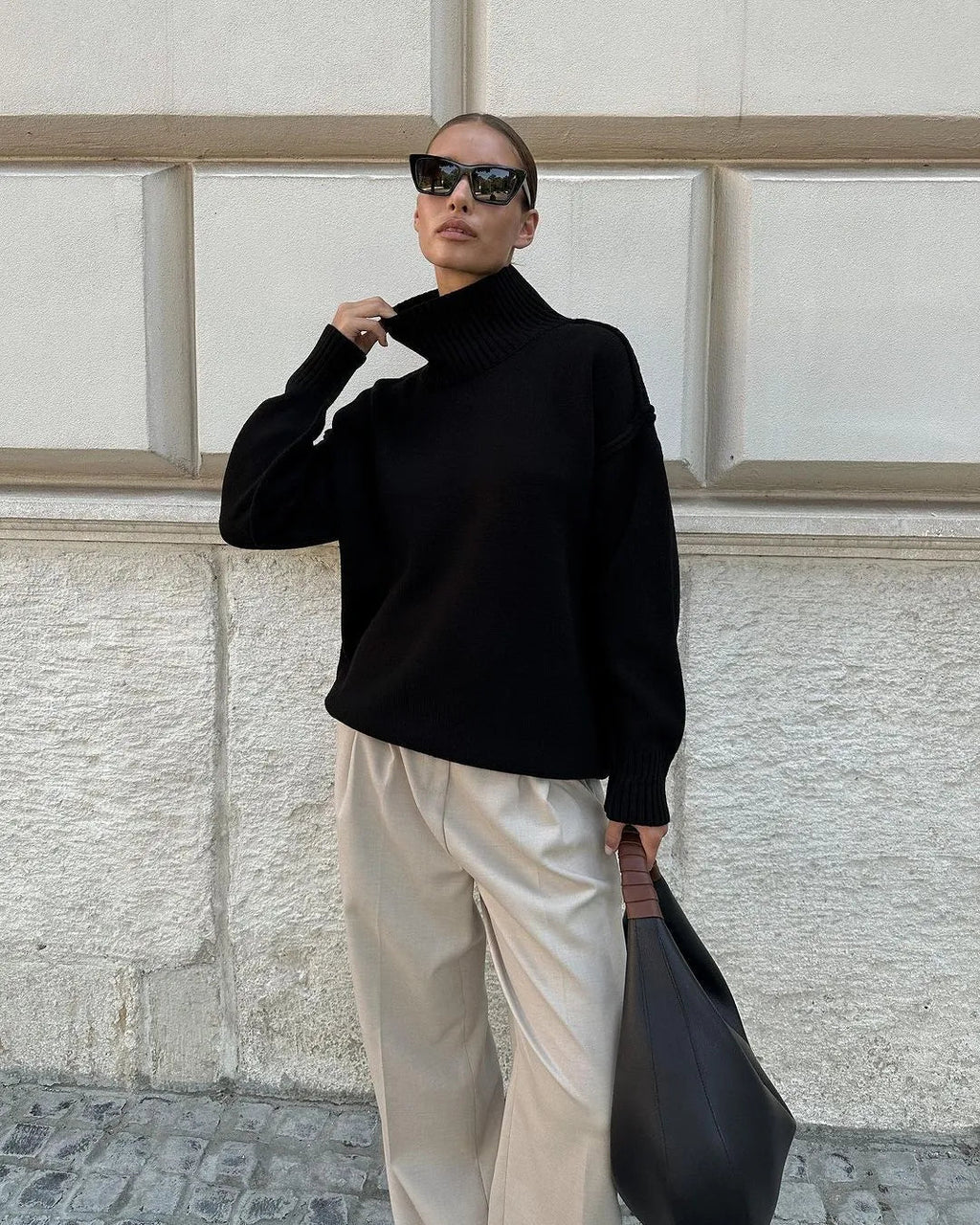 Femme élégante en pull noir à col roulé, pantalon beige ample, lunettes de soleil, sac à main noir, posant devant un mur texturé. Mode automne chic.