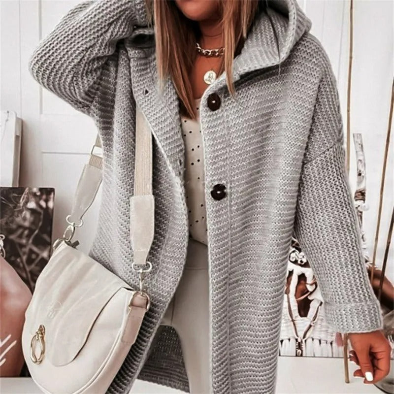 Femme portant un manteau en tricot gris avec capuche, boutons marron et sac à bandoulière blanc. Mode automne-hiver, style décontracté chic.