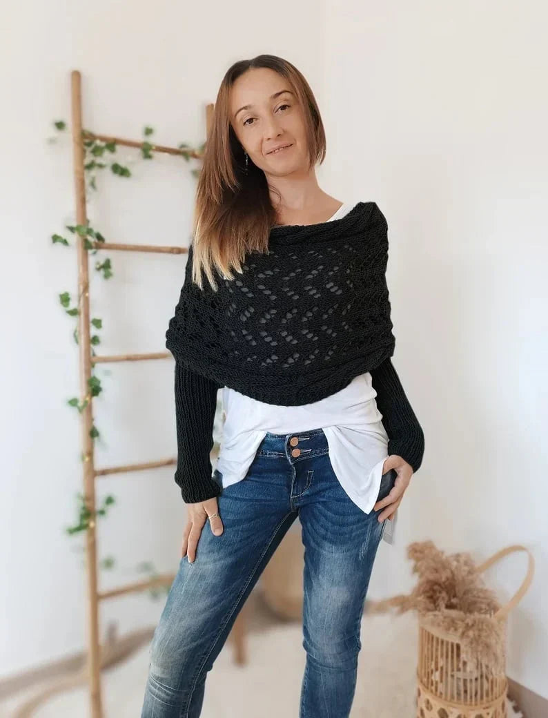 Femme portant un pull noir en maille ajourée, debout devant une échelle en bois décorée de plantes vertes. Mode décontractée, style automne-hiver.