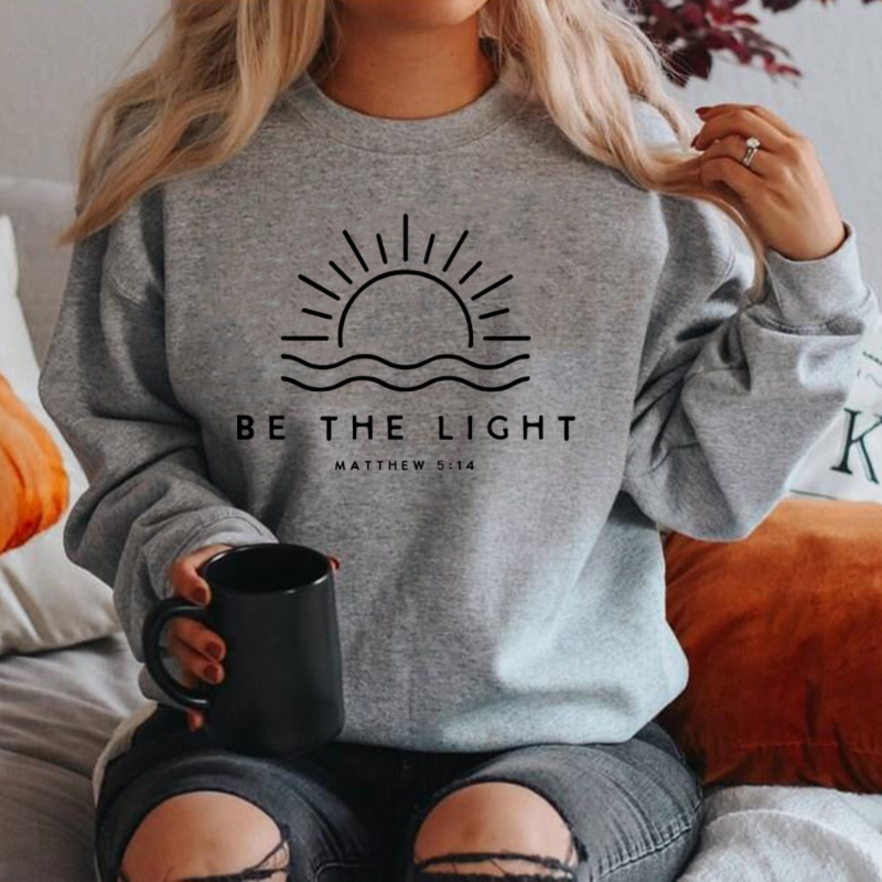 Femme portant un sweat gris avec un motif de soleil et l'inscription "Be the Light", tenant une tasse noire. Mode décontractée, style cosy.