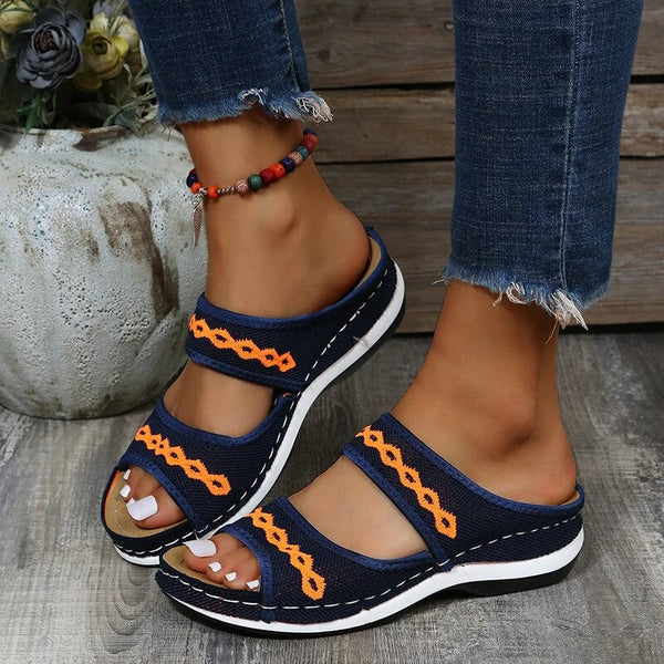 Sandales bleues à motifs orange sur pieds féminins, posées sur un sol en bois. Jean effiloché et bracelet perlé coloré. Mode estivale décontractée.