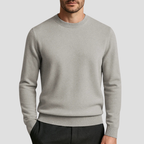 Homme portant un pull gris clair en laine, col rond, manches longues, style décontracté, sur fond uni. Mode masculine élégante et moderne.