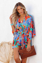 Femme en robe kimono à motifs floraux colorés, tenant une pochette marron, posant contre un mur blanc. Mode estivale, style bohème chic.