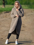Femme portant un long cardigan à capuche en tricot beige, motif torsadé, sur un chemin de terre, style décontracté, mode automne-hiver.