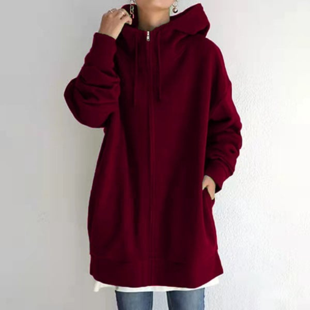 Personne portant un sweat à capuche rouge bordeaux ample, manches longues, avec fermeture éclair, sur fond blanc. Mode décontractée, tendance automne.