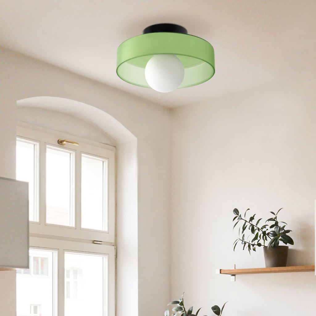 Plafonnier moderne vert avec abat-jour circulaire, installé dans un salon lumineux avec plantes et étagère en bois. Éclairage intérieur design.