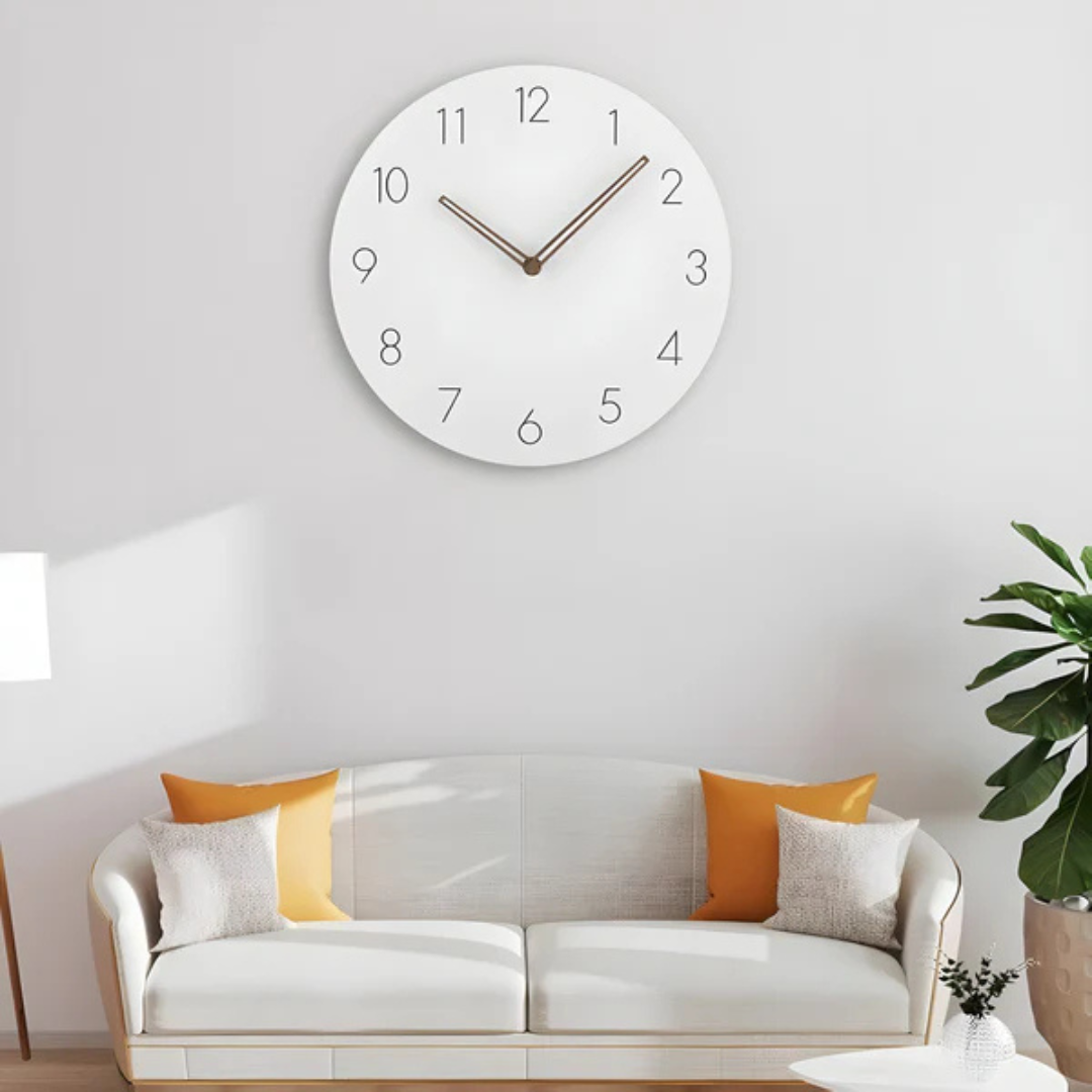 Horloge murale moderne blanche avec chiffres noirs, au-dessus d'un canapé blanc avec coussins orange, dans un salon lumineux et minimaliste.