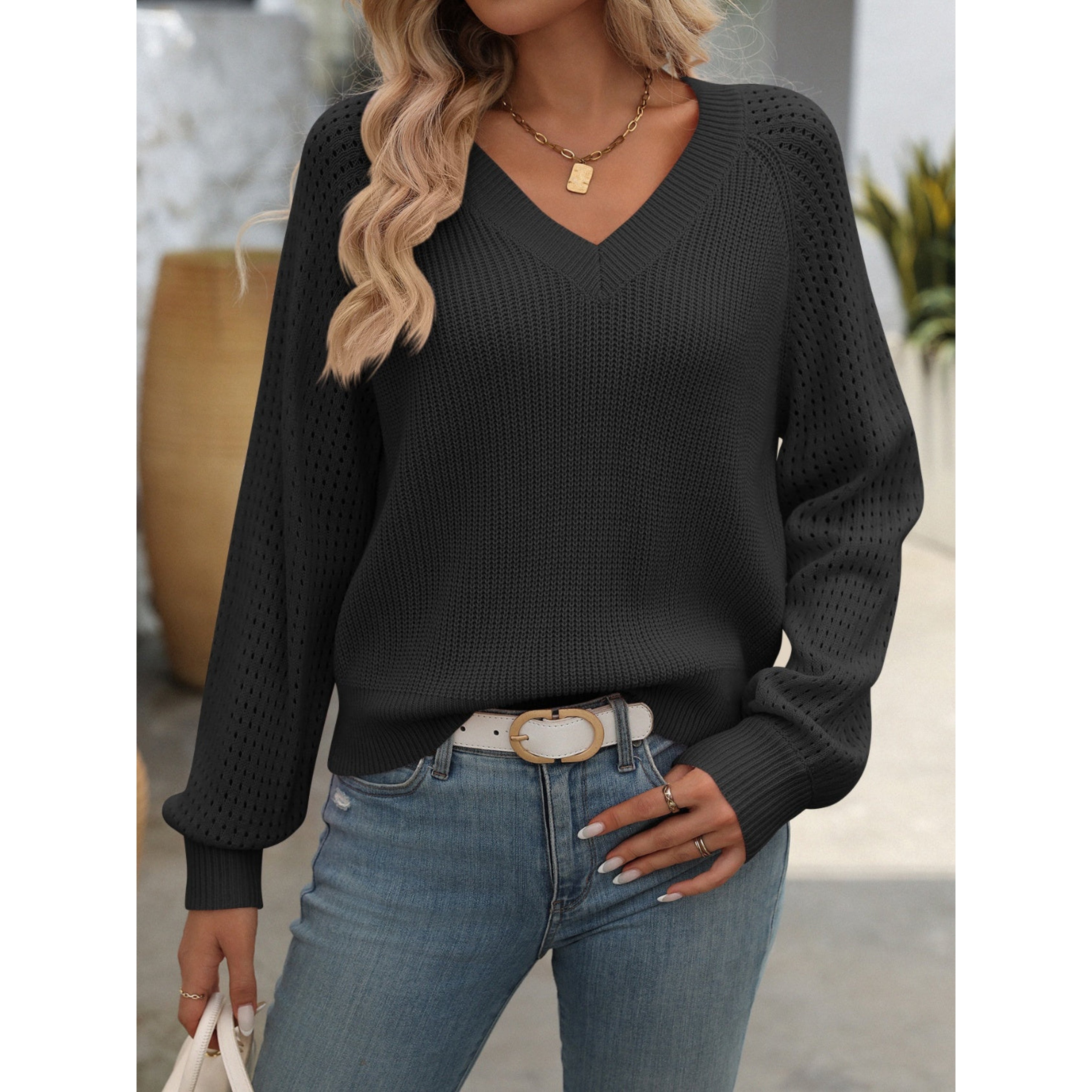 Femme portant un pull noir à col en V, manches longues, avec un jean bleu et une ceinture blanche, accessoirisée d'un collier doré.