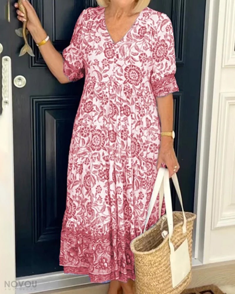 Robe longue à motifs floraux rouges et blancs, femme tenant un sac en osier devant une porte noire. Mode estivale élégante et décontractée.