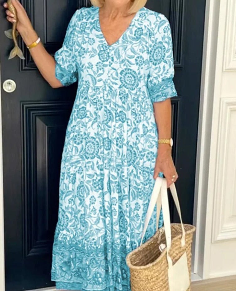 Femme en robe longue bleue à motifs floraux, tenant un sac en osier, devant une porte noire. Mode estivale élégante et décontractée.